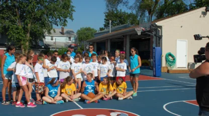 A group of children and adults pose for a photo on an outdoor basketball court, with some kids in white shirts and others in blue and yellow shirts. A camera operator films them.