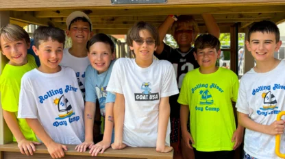 A group of eight boys stand and smile inside a wooden playhouse at day camp, wearing shirts labeled "Rolling River Day Camp" and "Goat USA.