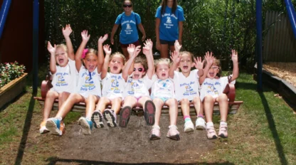 A group of young children sit on a large swing with their hands raised, smiling, while two adults stand behind them outdoors on a sunny day.