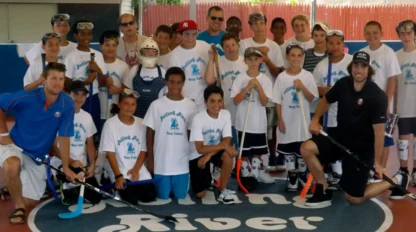 A group of children and adults wearing matching t-shirts pose with hockey sticks on an outdoor court, some kneeling and some standing, at a hockey camp.