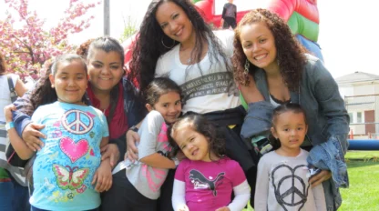 Three women and four young girls pose and smile outdoors near an inflatable slide on a sunny day, with trees and a building visible in the background.