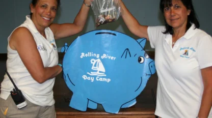 Two women stand on either side of a large blue piggy bank labeled "Rolling River Day Camp," holding up a plastic bag of coins together.