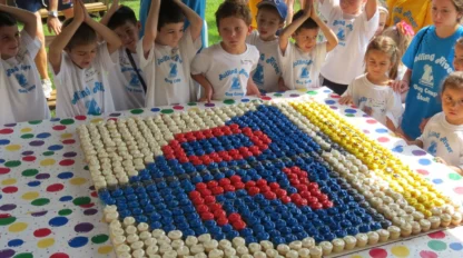 Children in matching t-shirts stand around a large table of cupcakes arranged to form a colorful logo, as they pose with hands above their heads.