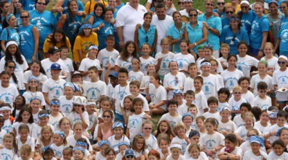 A large group of children and adults pose outdoors for a photo, most wearing matching camp shirts in white or teal, with trees and playground equipment in the background.