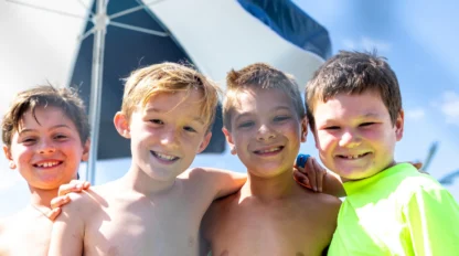 Four young boys stand close together outdoors, smiling at the camera. An umbrella and blue sky are visible in the background.