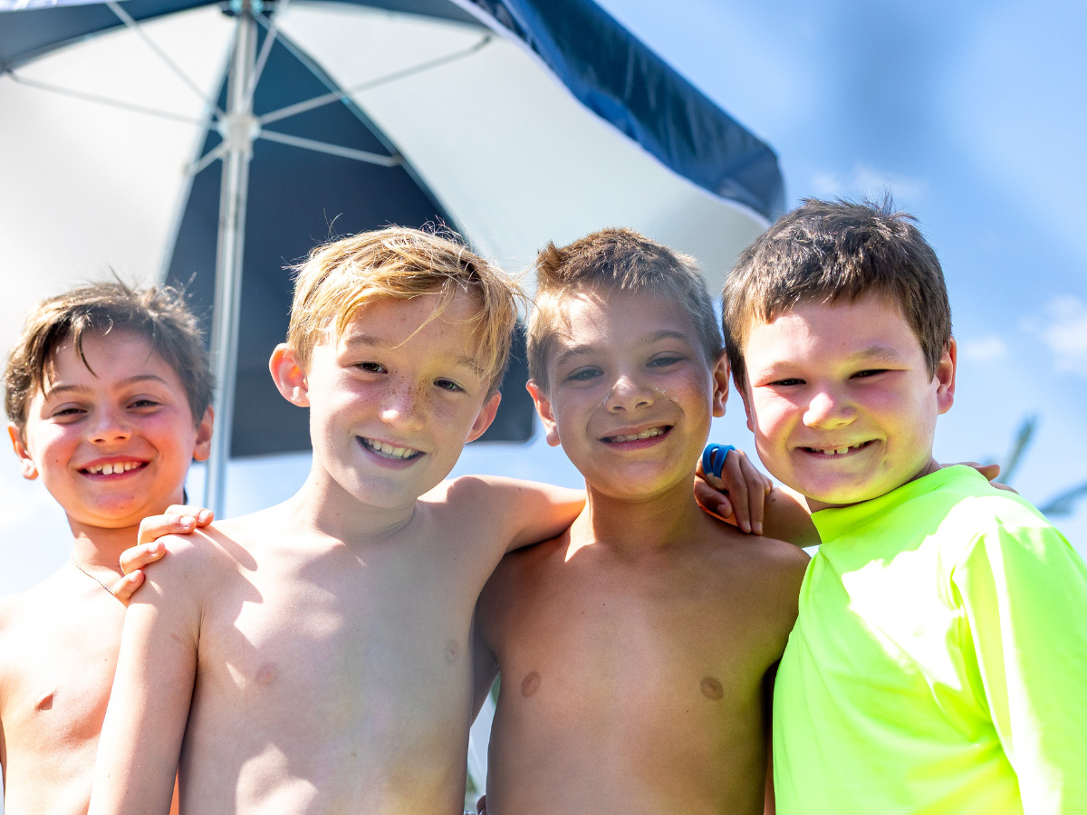 Four young boys stand close together outdoors, smiling at the camera. An umbrella and blue sky are visible in the background.