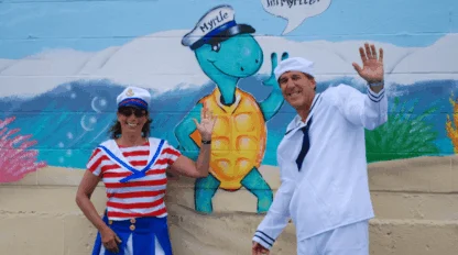 Two adults dressed in sailor costumes stand in front of a mural featuring a turtle in a sailor hat that says, "Myrtle" and a speech bubble reading, "Hi kids, I'm Myrtle.