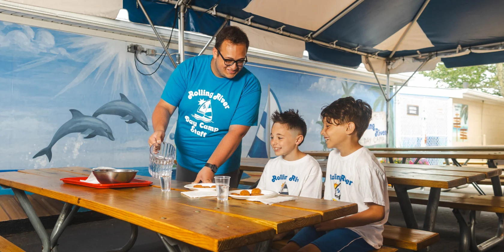 An adult pours water into a glass for two boys sitting at a picnic table with food trays, under a canopy with a mural of dolphins in the background.