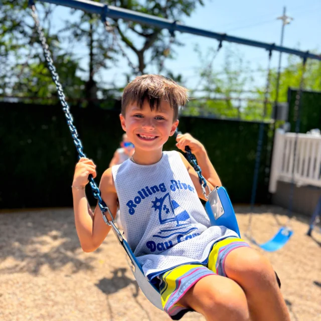 A young boy in a white "Rolling River Day Camp" shirt smiles while sitting on a swing at an outdoor playground on a sunny day.