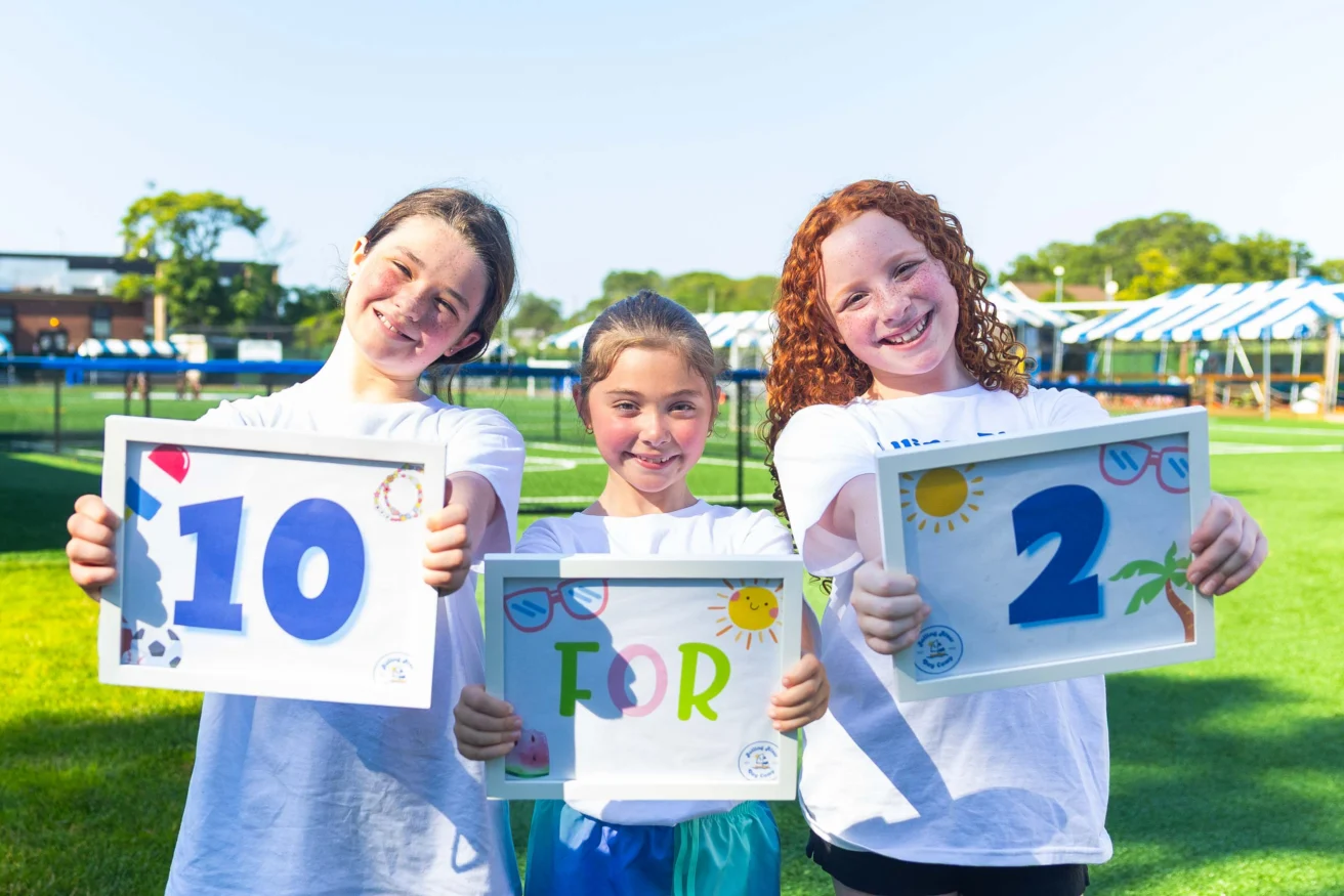 Three children standing outdoors on grass hold up signs reading “10 FOR 2” while smiling at the camera.