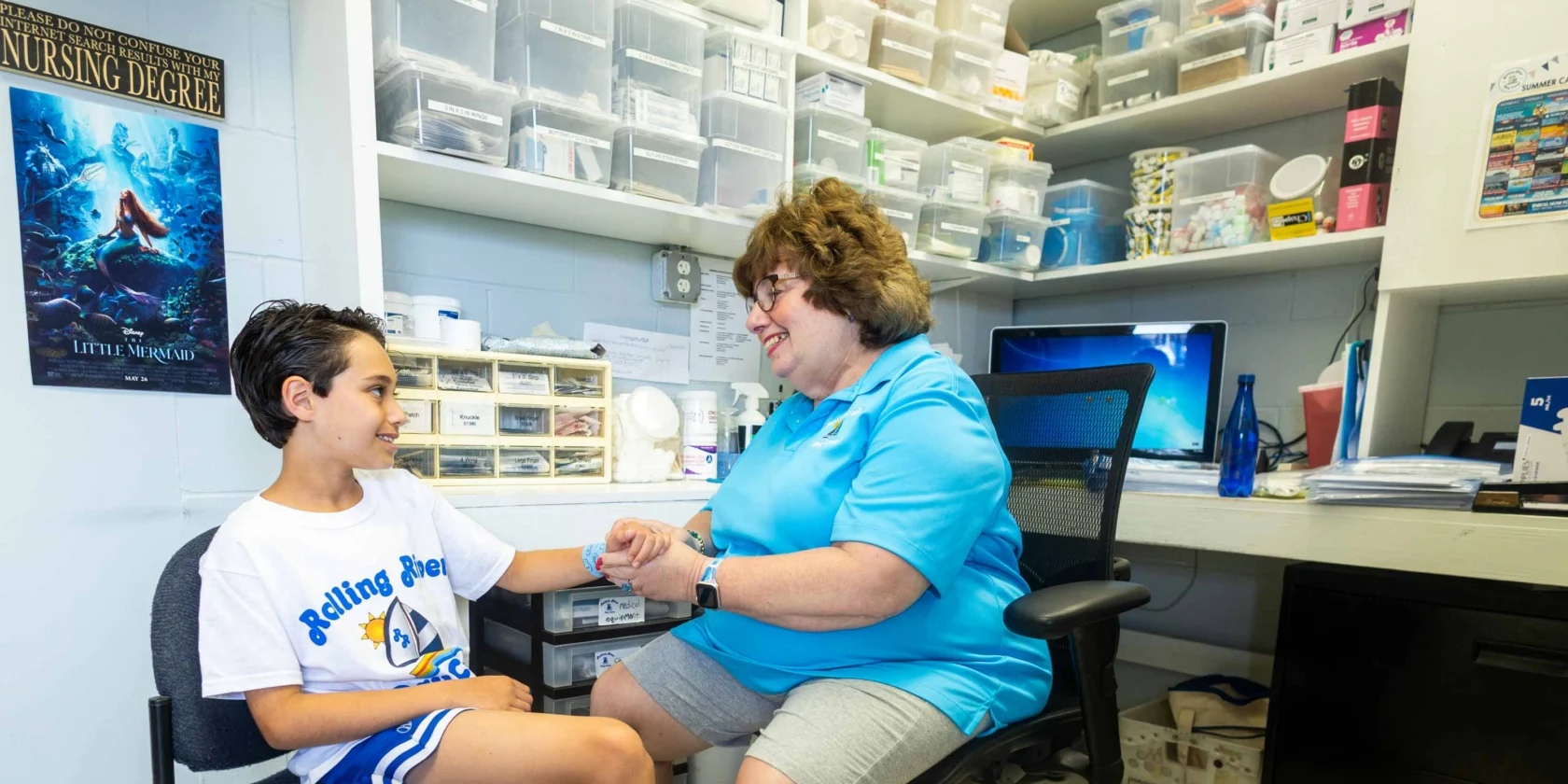 A woman in a blue shirt talks with a boy in a Rolling Stones t-shirt in an office or medical room filled with organized supplies.