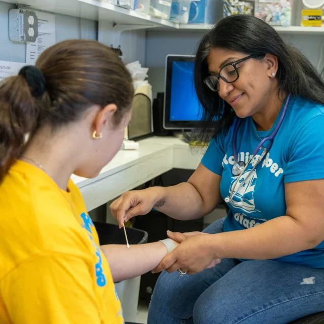 A healthcare professional examines a child's arm with a tongue depressor in a medical office. Both are wearing yellow shirts, and various medical supplies are visible in the background.