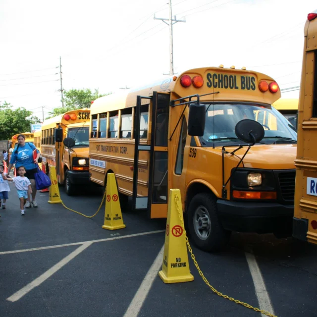 Yellow school buses are parked with "NO PARKING" cones in front, as children and adults walk nearby on a sunny day.