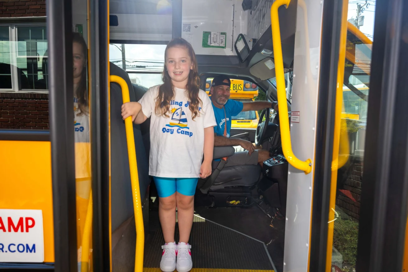A young girl stands at the entrance of a school bus, smiling at the camera, while a bus driver sits at the driver's seat and looks toward her.