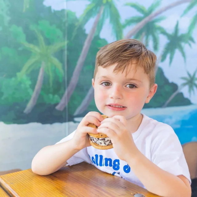 A young boy sits at a wooden table holding an ice cream sandwich, with a tropical beach mural in the background.