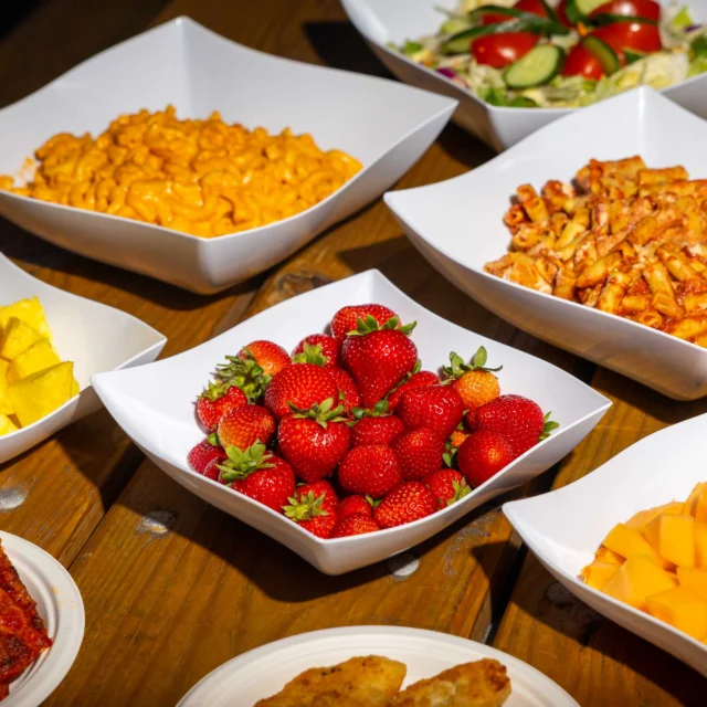 Assorted dishes on a wooden table, including macaroni and cheese, baked pasta, salad, sliced pineapple, strawberries, mango, pizza, and breaded food.