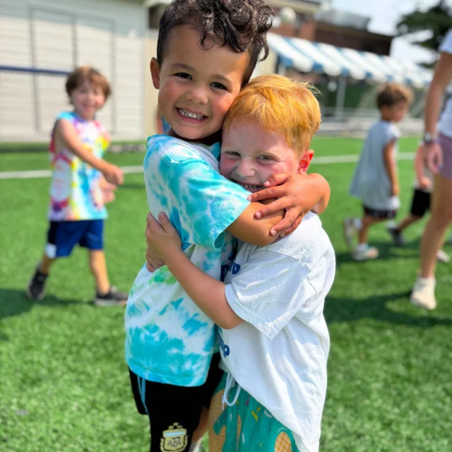 Two young boys stand on a grassy field, smiling and hugging each other. Other children are visible playing in the background.