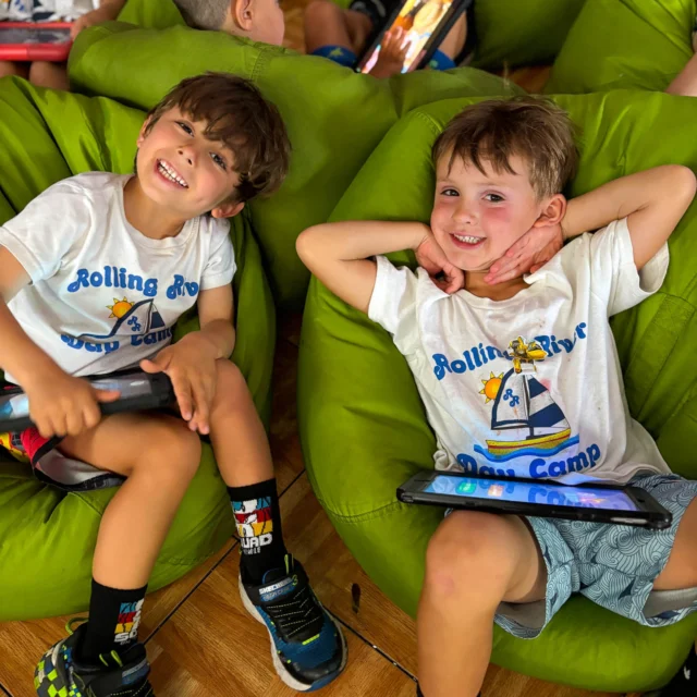Two young children sit on green bean bags, smiling and holding tablets, wearing matching "Rolling River Day Camp" t-shirts. Other children are visible in the background.