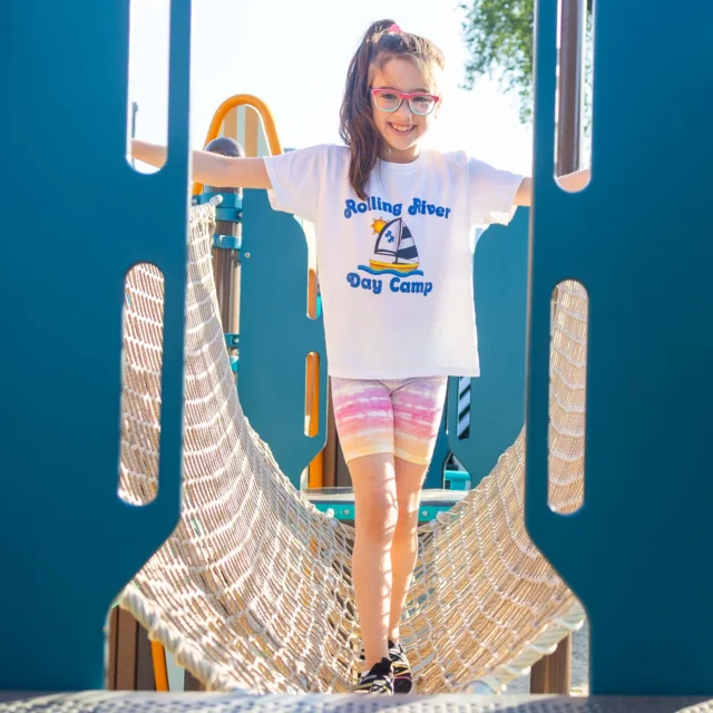 A girl wearing a "Rolling River Day Camp" shirt and glasses stands on a rope bridge at a playground, smiling at the camera.