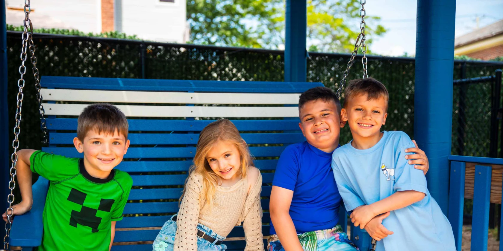 Four children sit and smile on a blue swing bench outdoors, dressed in summer clothes, with trees and a fence in the background.