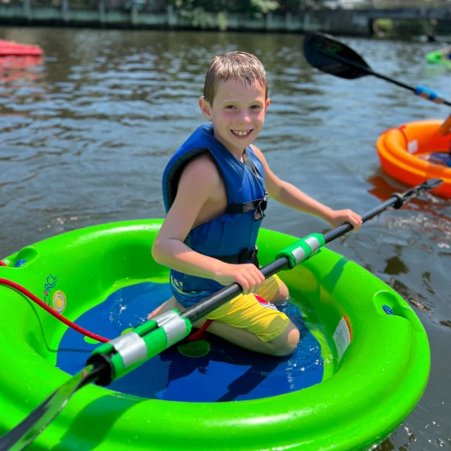 A boy wearing a blue life jacket sits in a green circular boat on a sunny day, paddling on a calm river.