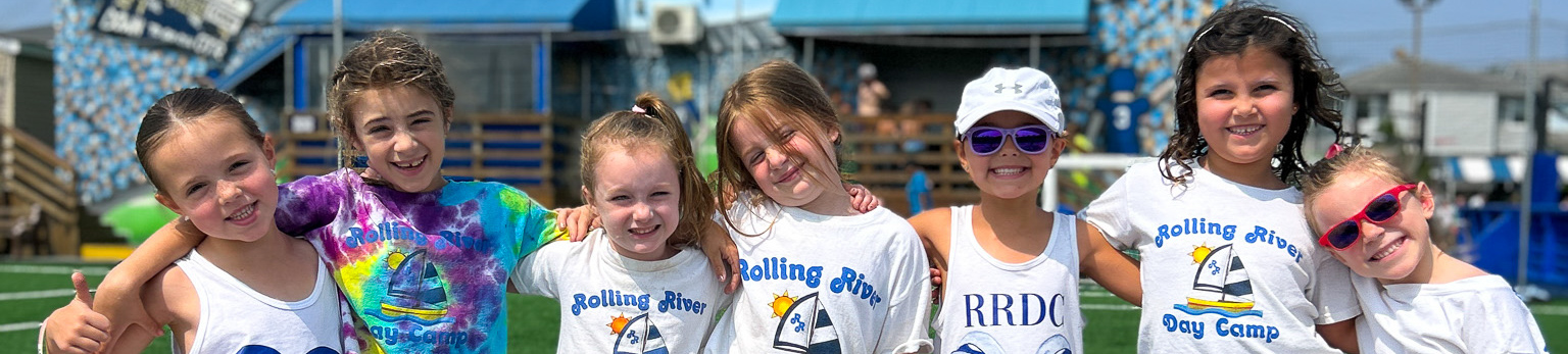 Six young girls stand in a row on a sports field, smiling at the camera, wearing camp shirts and summer outfits, with a building in the background.
