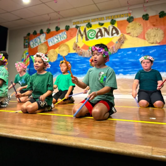 Children dressed in green shirts and flower crowns perform with sticks on stage in front of a "Moana" themed backdrop at a school event.