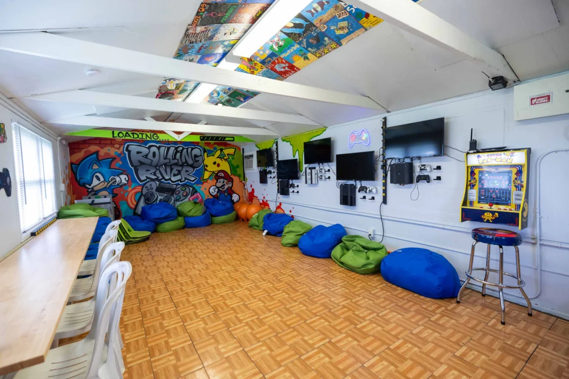 A game room with bean bags, wall-mounted TVs, video game consoles, an arcade machine, and colorful graffiti art on the walls.