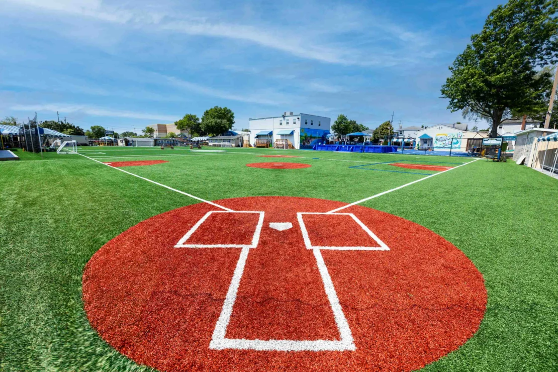 A baseball field with artificial turf shows the home plate area and bases, surrounded by green grass and a few buildings and trees in the background under a blue sky.