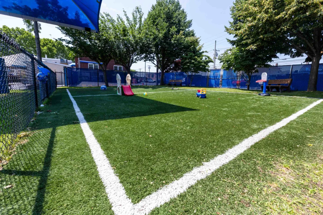 A fenced outdoor play area with artificial turf, white boundary lines, playground equipment, benches, and trees providing shade.