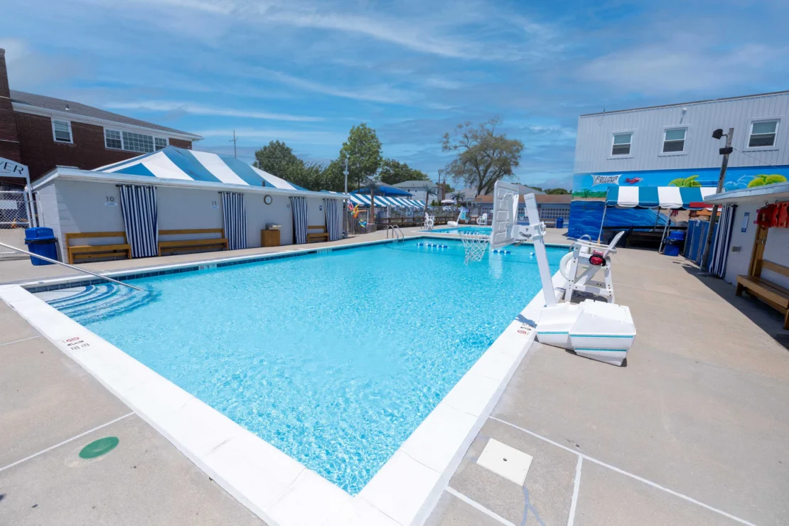 Outdoor swimming pool surrounded by benches, cabanas, and buildings, with a pool lift and basketball hoop on the pool deck under a clear sky.
