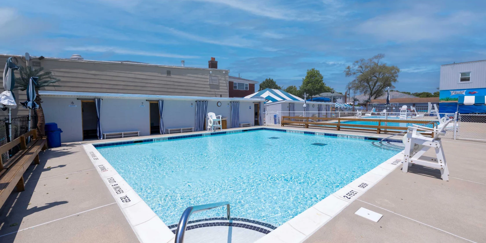 Outdoor swimming pool with clear water, surrounded by lounge chairs, cabanas, and safety equipment on a sunny day.