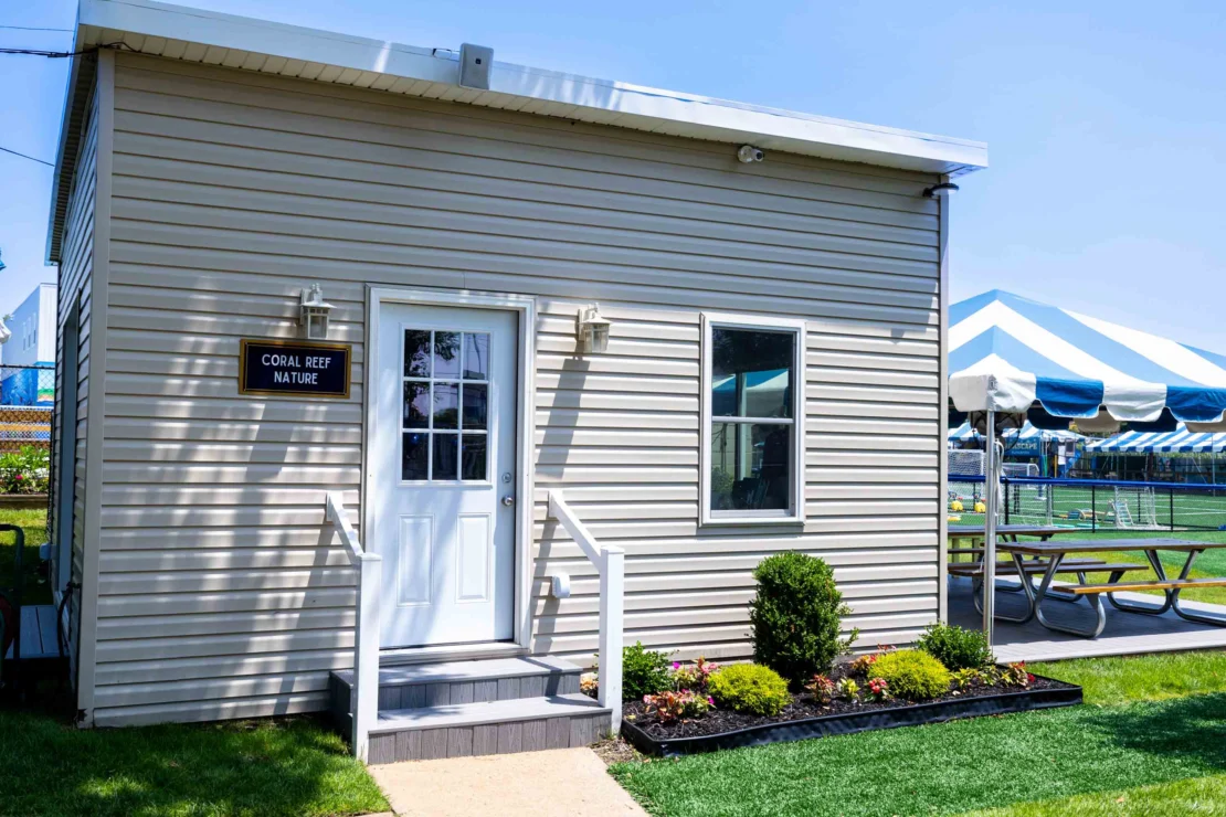 Small beige modular building with a sign reading "Coral Reef Nature," steps leading to a white door, plants in front, and a striped tent with picnic tables in the background.