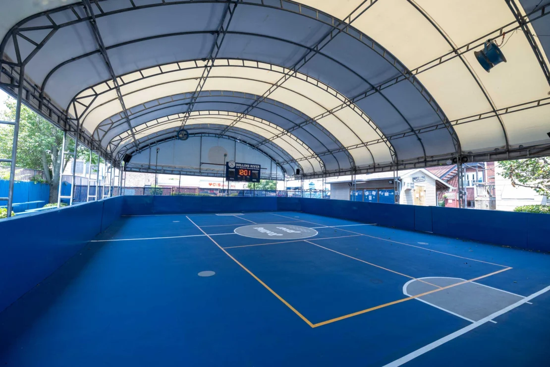 Covered blue futsal court with white and yellow markings, scoreboard at one end, surrounded by partial blue walls and natural light coming in from the sides.
