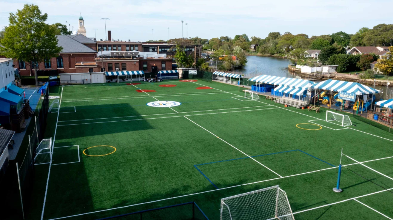 Aerial view of an empty artificial turf sports field with soccer goals, bordered by tents and buildings, situated near a river.