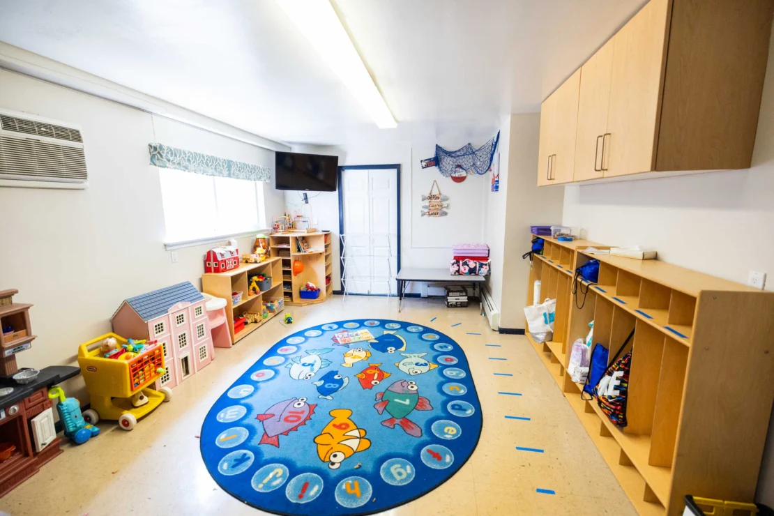 A brightly lit preschool classroom with a blue fish-themed rug, shelves of toys, cubbies with bags, and wall cabinets lining the room.