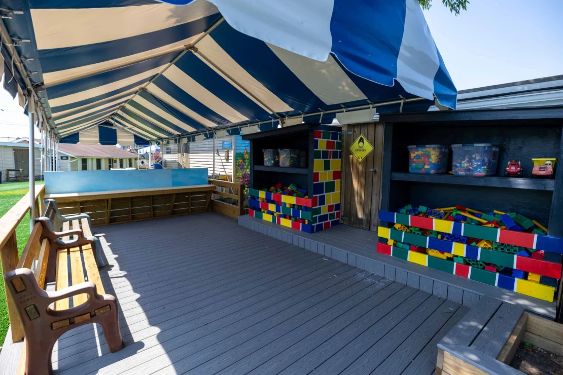 Outdoor play area with benches, storage shelves, and large colorful building blocks under a blue and white striped canopy.