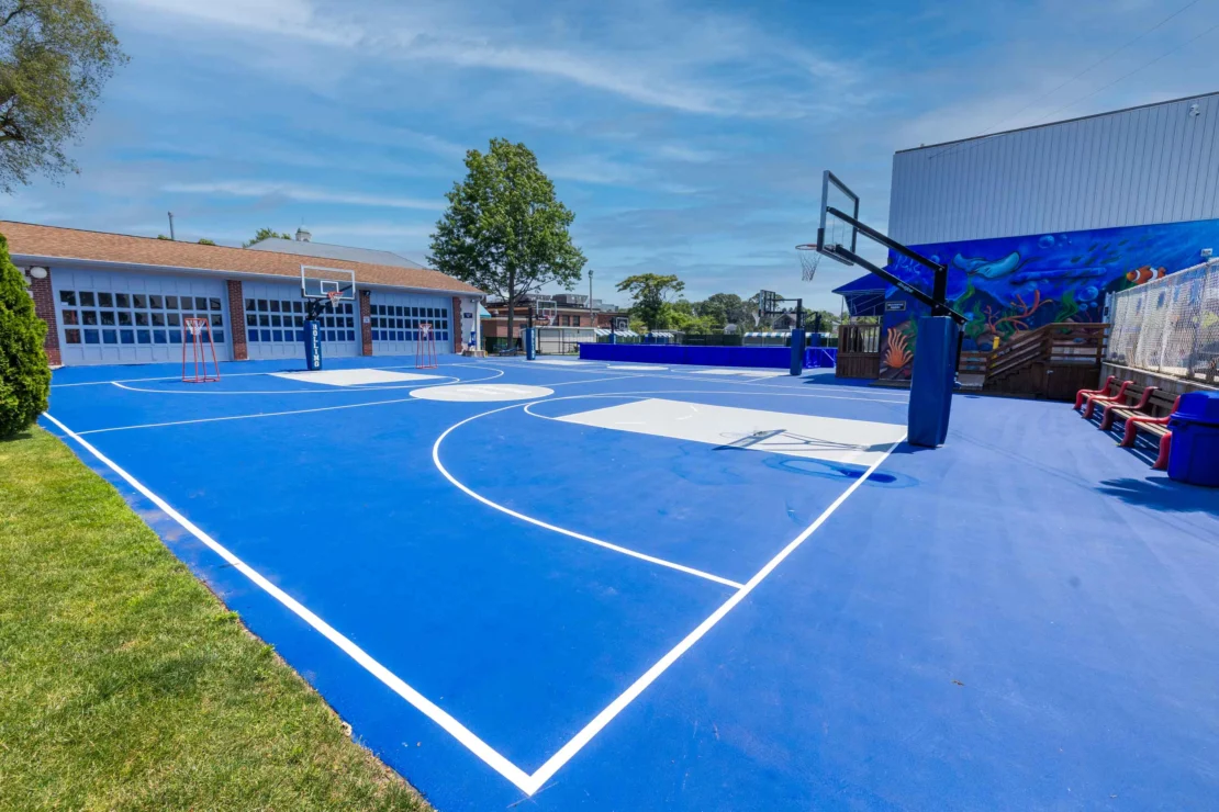 Outdoor blue basketball court with two hoops, white lines, and benches on the side under a clear sky. Building and mural are visible in the background.