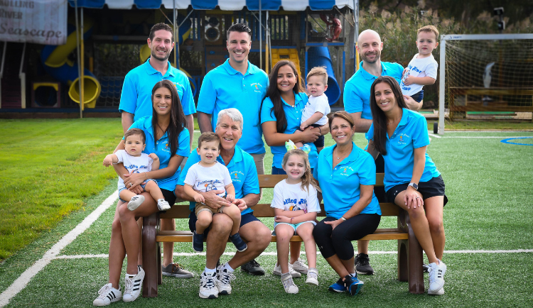 A group of adults and children wearing matching blue shirts pose for a photo on a soccer field with playground equipment in the background.