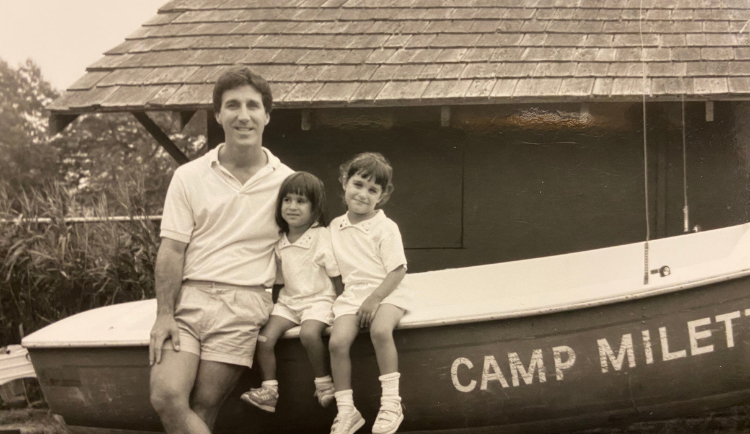 A man and two children pose in front of a small boat labeled "CAMP MILET" with a shingled-roof building in the background. All three wear light-colored shirts and shorts.