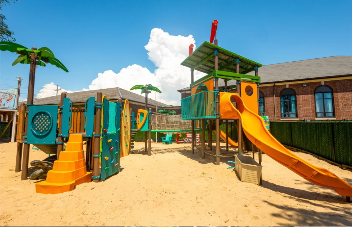 Outdoor playground with yellow slides, climbing structures, fake palm trees, sand surface, and colorful chairs, situated next to a brick building under a clear blue sky.