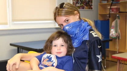 A camp counselor wearing a mask, hugging a camper.