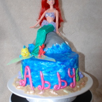 Ariel Cake Chocolate cream cake covered with buttercream frosting that has been hand painted blue, gum paste letters, fondant seashells and sea weed,...