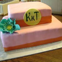 Colorful Monogram Cake Two tier cake covered in pink vanilla fondant. Orange fondant borders. Teal tinted gumpaste flower. Yellow gumpaste plaque with monogram...