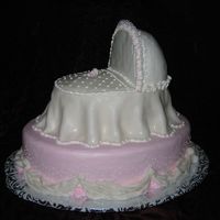 Baby Mine Baby bassinet with soft fondant drapes and royal icing fringe around the top of the hood. A small baby rattle lays on the quilt. The pink...