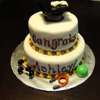 Exercise And Sports Science Theme Grad Cake Two tier cake covered in white fondant with maroon and gold piped edges. The topper is a grad cap made of cake, covered in fondant and...