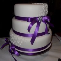 Three Tier Round Wedding Cake With Purple Ribbon And Brooch Detail Cake iced in fondant and has blossom and petal detail with two vintage look brooches.