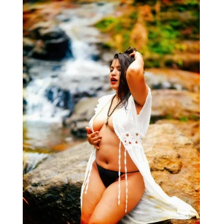 Anjali Gaud with pale full breasts and black thong wearing open white lace robe with beaded necklace near waterfall in natural landscape