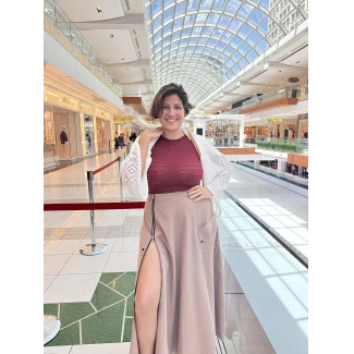 Woman with large full breasts in maroon form-fitting top and light brown high-waisted skirt with thigh-high slit posing in shopping mall with luxury storefronts visible