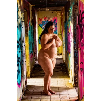 Full-figured woman with large pendulous breasts and light pink nipples wearing pearl necklace standing in graffiti-covered hallway with dramatic sunlight